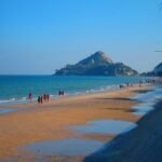 Plage de Hua Hin avec promeneurs sur le sable, mer turquoise et colline rocheuse surmontée d'un temple en arrière-plan