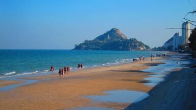 Plage de Hua Hin avec promeneurs sur le sable, mer turquoise et colline rocheuse surmontée d'un temple en arrière-plan